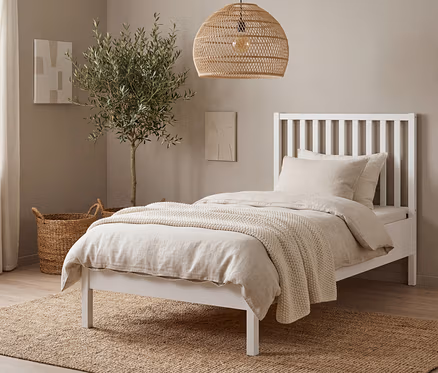 Ein weißes Bett mit beiger Bettwäsche steht auf einem braunen Teppich. Eine Korblampe hängt über dem Bett, neben dem ein Olivenbaum steht.