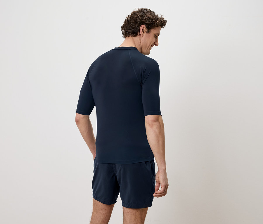 Mann von hinten in marineblauem Shirt mit kurzen Ärmeln und Shorts vor einem weißen Hintergrund.