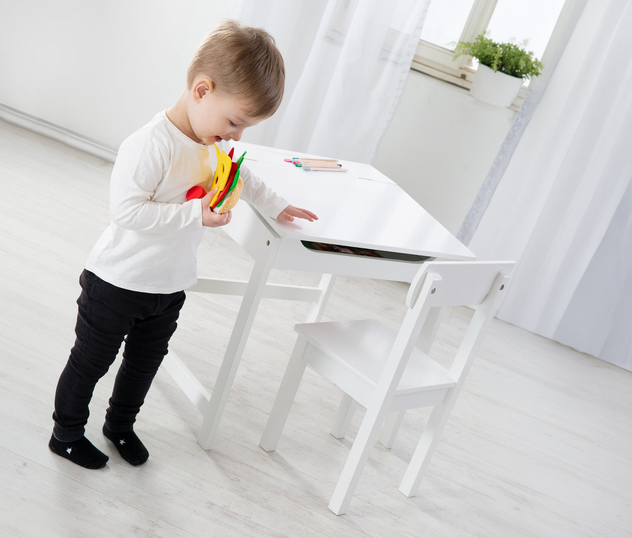 Kleiner Junge mit Spielzeug-Sandwich steht an einem weißen Tisch mit Stuhl. Buntstifte liegen auf dem Tisch.