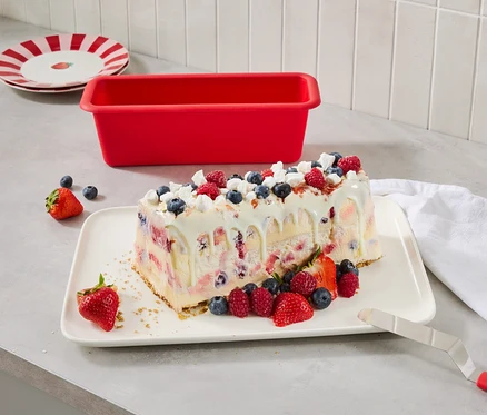 Eisdessert mit Beeren auf einem Teller, daneben eine rote Kuchenform und Teller mit roten Streifen.