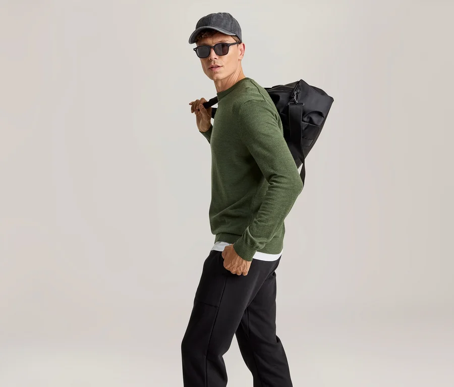 Mann geht, blickt über die Schulter, trägt grünes Langarmshirt, schwarze Jogginghose, graue Baseballkappe, schwarze Sonnenbrille und eine schwarze Sporttasche über der Schulter.