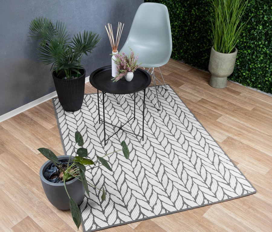 Grau-weißer Outdoor-Wendeteppich »Knitted Joy« mit geflochtenem Muster auf Holzboden, umgeben von einem schwarzen Beistelltisch, einem hellblauen Stuhl und mehreren Topfpflanzen.