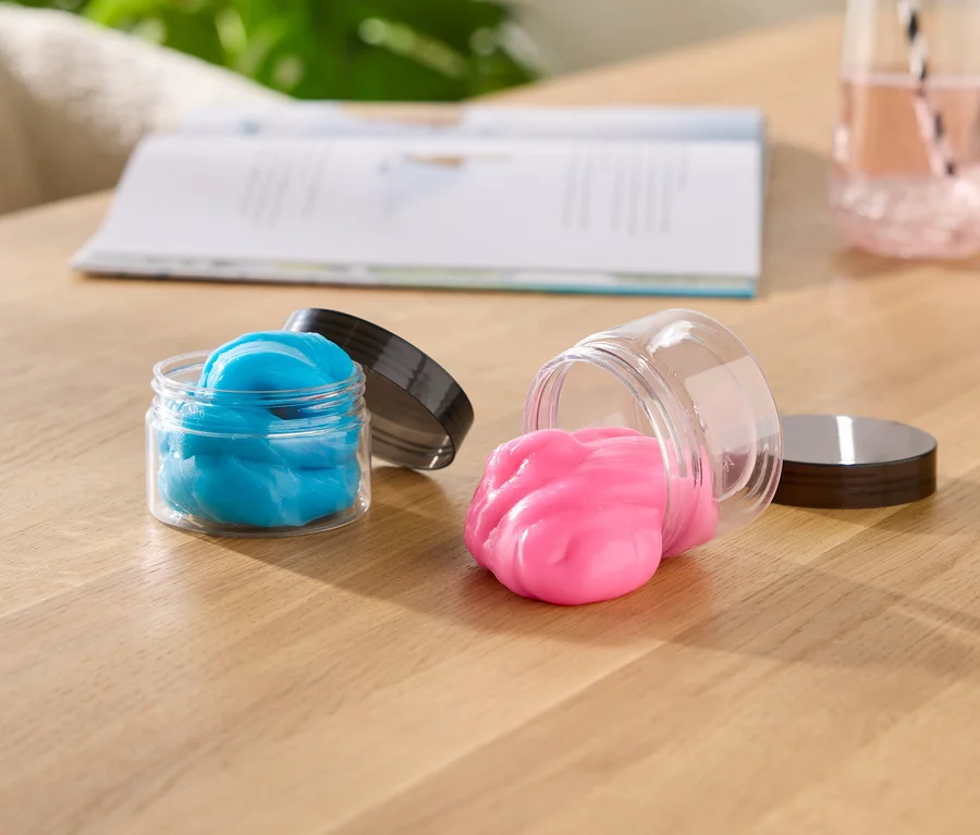Zwei offene Schleimbehälter auf einem Holztisch; einer aufrecht mit blauem Schleim, der andere liegt seitlich, wobei pinker Schleim herausquillt. Ein offenes Buch und ein Glas mit rosa Flüssigkeit sind im Hintergrund.