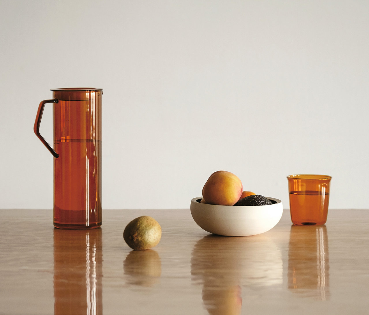KINTO Krug »Cast Amber« mit Wasser auf einem Holztisch, daneben eine Limette, eine Schale mit Pfirsichen und Avocados sowie ein bernsteinfarbenes Glas Wasser.