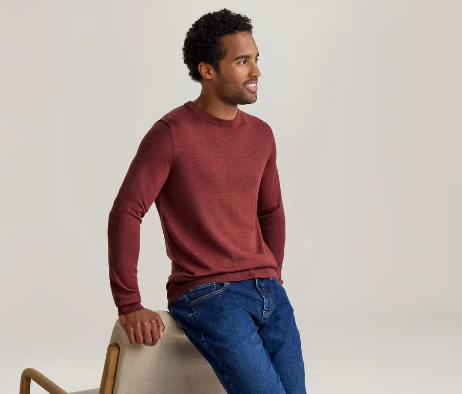 Mann in einem roten Langarmshirt und blauen Jeans, der sich lächelnd an einen hellen Stuhl lehnt und nach rechts blickt.