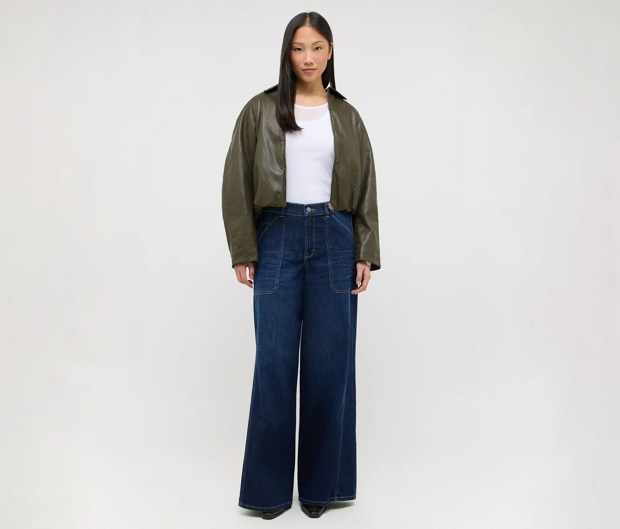 Eine Frau steht frontal, trägt ein weißes T-Shirt, eine kurze olivgrüne Jacke und eine dunkelblaue weite Jeans.
