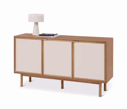 Helles Sideboard aus Holz mit drei cremefarbenen Türverkleidungen und vier Beinen. Darauf stehen eine weiße Lampe und ein Stapel Zeitschriften.