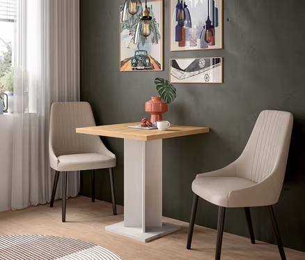 Ein kleiner Essbereich mit einem quadratischen Holztisch, zwei beigefarbenen Polsterstühlen und drei gerahmten Kunstwerken an einer dunkelgrünen Wand. Zwei Pendelleuchten hängen über dem Tisch, auf dem eine rosa Vase, eine weiße Tasse und Macarons stehen. Ein runder Teppich liegt auf dem Holzboden.