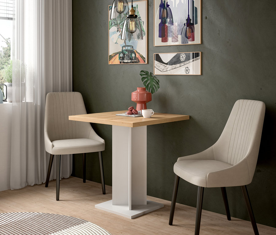 Ein kleiner Essbereich mit einem quadratischen Holztisch, zwei beigefarbenen Polsterstühlen und drei gerahmten Kunstwerken an einer dunkelgrünen Wand. Zwei Pendelleuchten hängen über dem Tisch, auf dem eine rosa Vase, eine weiße Tasse und Macarons stehen. Ein runder Teppich liegt auf dem Holzboden.