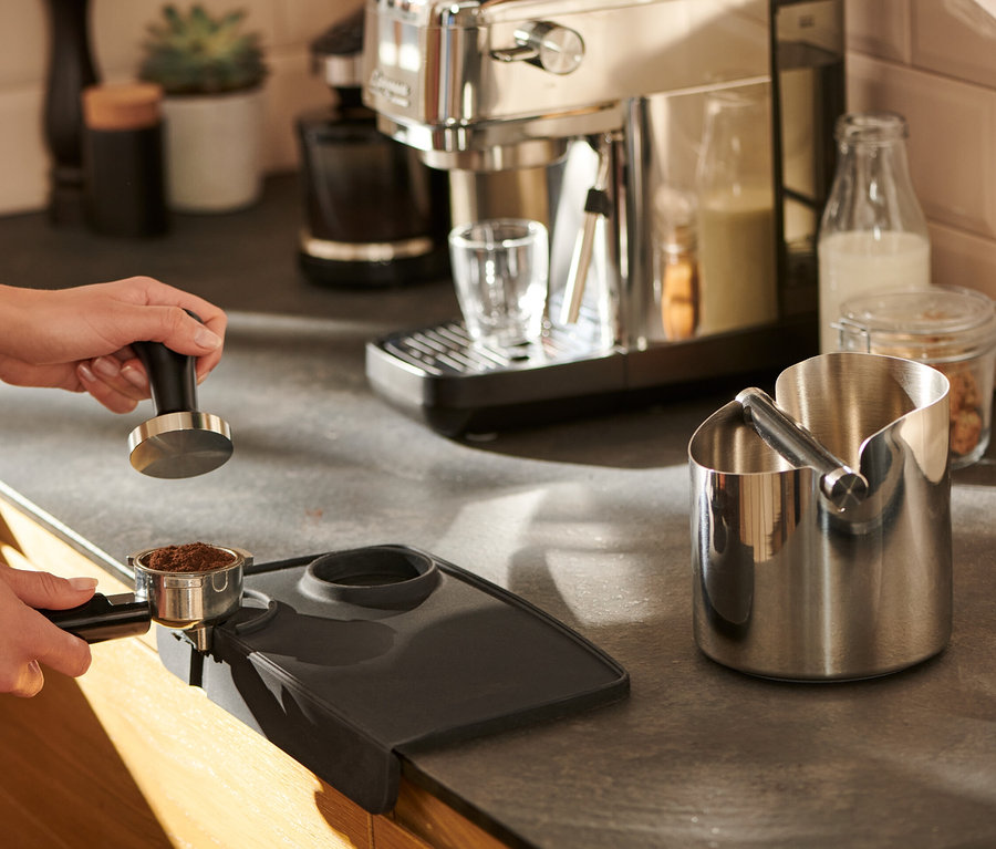 Eine Frau tampert Kaffee auf einer schwarzen Tampermatte.