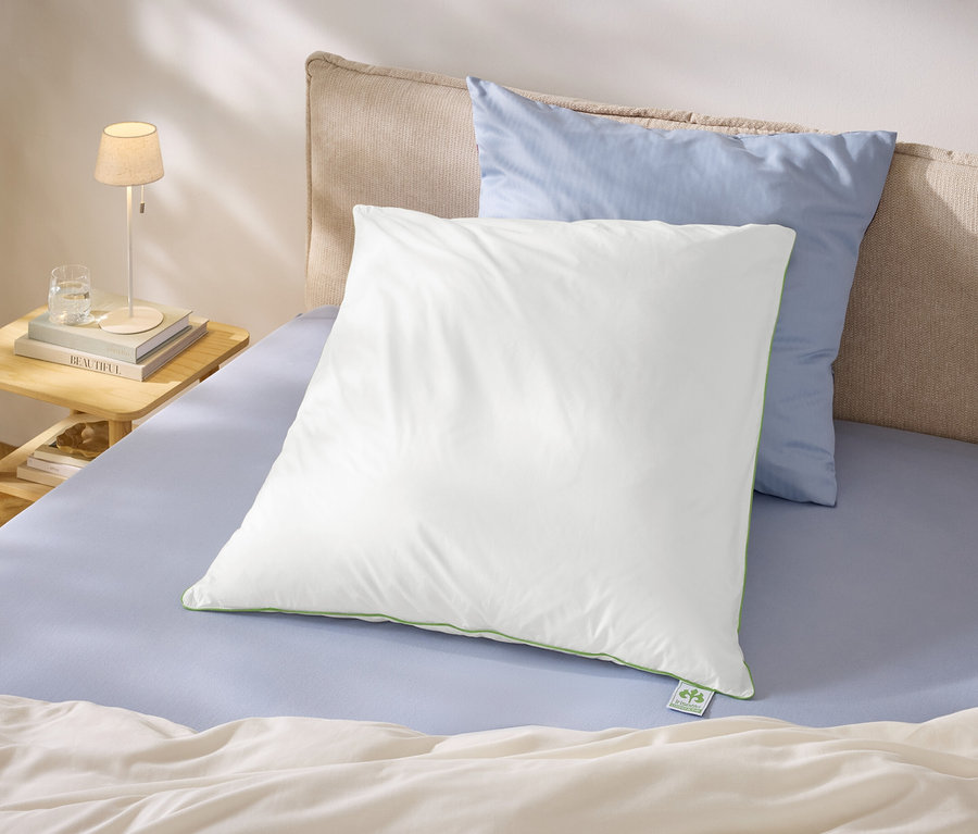 Bett mit hellblauem Spannbettlaken, einem großen weißen Kissen mit grüner Paspelierung und einem hellblauen Kissen. Daneben ein Nachttisch mit Lampe, Büchern und Wasserglas.