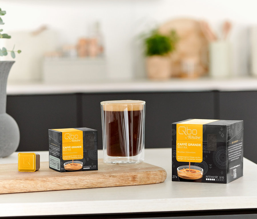 Ein Glas Kaffee steht neben einer Qbo Caffè Grande BELO SUL - 8 Kapsel Packung und einer einzelnen Kapsel auf einem Holzbrett.