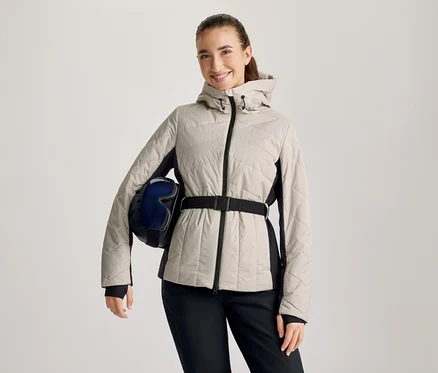 Lächelnde Frau in beiger Steppjacke mit schwarzen Seitenpartien und schwarzem Gürtel, schwarzer Hose, hält einen dunkelblauen Skihelm mit Skibrille unter dem linken Arm.