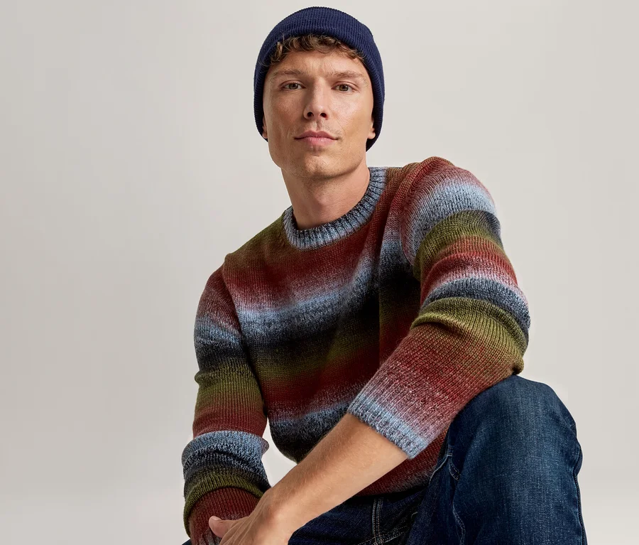 Mann trägt eine dunkelblaue Mütze, einen mehrfarbigen gestreiften Pullover in Blau-, Rot-, Grün- und Schwarztönen sowie blaue Jeans, sitzt und blickt nach vorne.