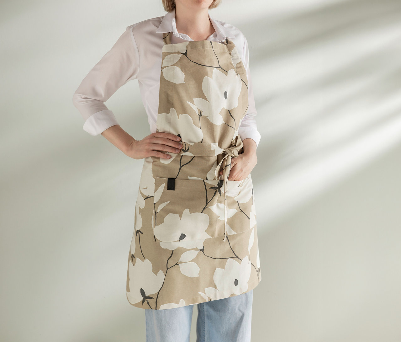 Frau trägt eine beigefarbene Schürze mit weißem Blumenmuster, weißes Hemd und blaue Jeans, steht mit den Händen in den Hüften.