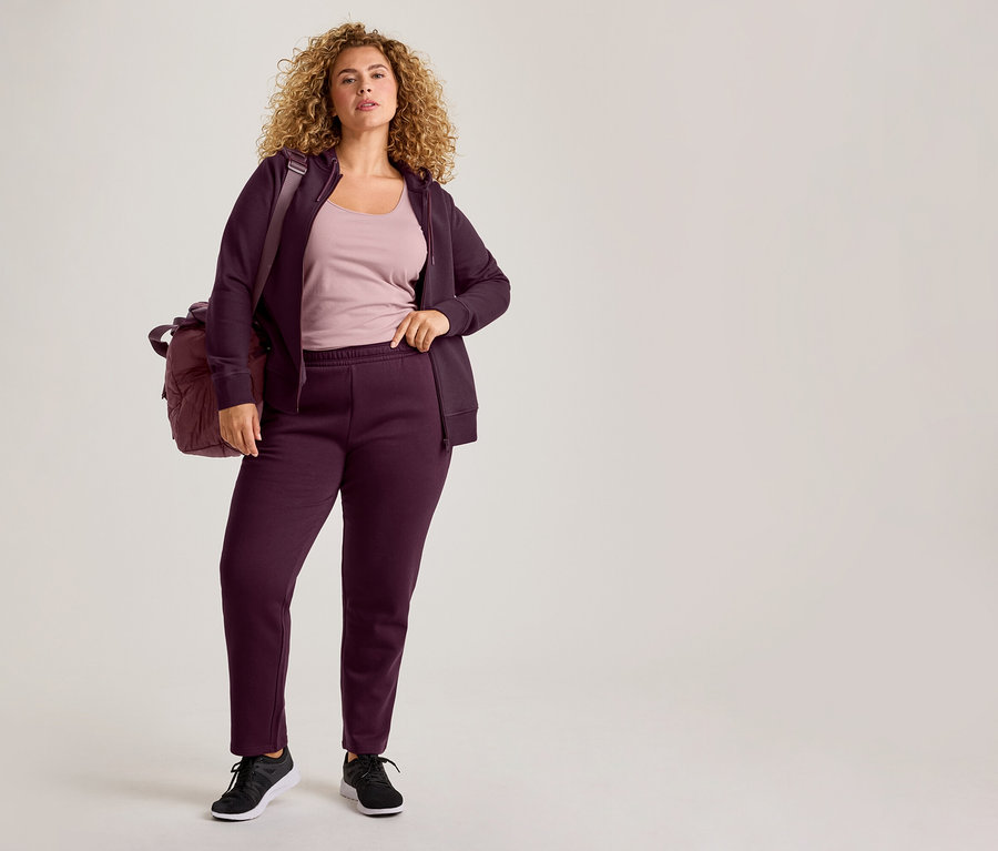 Eine Frau mit lockigem Haar trägt einen lila Kapuzenpullover mit Reißverschluss, passende Jogginghosen, ein hellrosa Tanktop und schwarze Turnschuhe, mit einer lila Sporttasche über der Schulter.