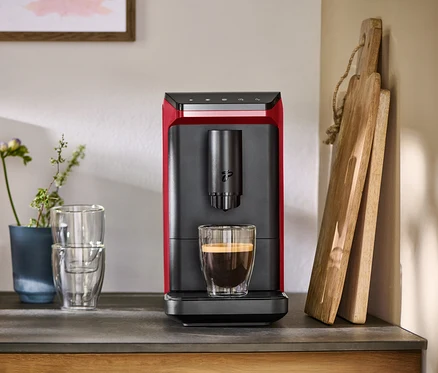 Roter und schwarzer Kaffeevollautomat mit einer Tasse Kaffee auf einer Arbeitsplatte, daneben Gläser und Schneidebretter.