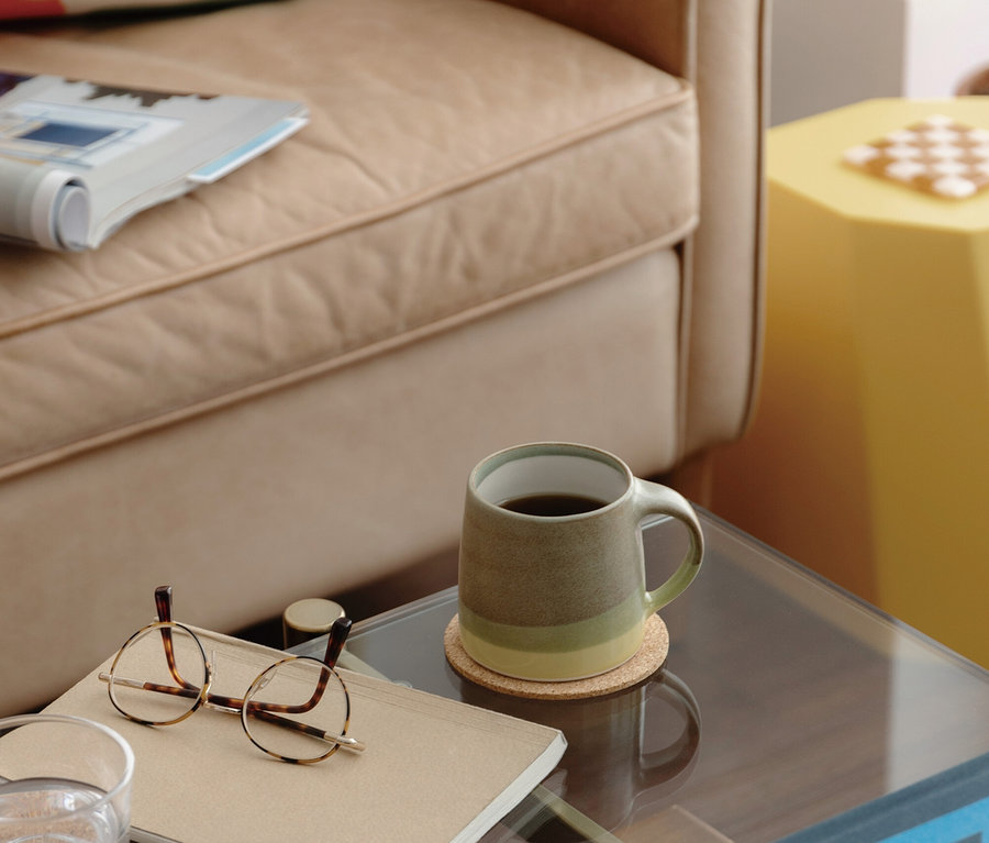 Ein beiger Sessel mit einem bunten Kissen und einer Zeitschrift steht neben einem Couchtisch aus Glas. Auf dem Tisch befinden sich eine grün-beige Kaffeetasse, ein Glas Wasser, eine Brille auf einem Notizbuch und ein Stapel Bücher. Im Hintergrund steht ein gelber, sechseckiger Beistelltisch.