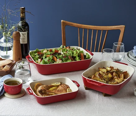 Drei kirschrote STAUB Auflaufformen auf einem Tisch, gefüllt mit Salat, Hähnchen und Kartoffeln.