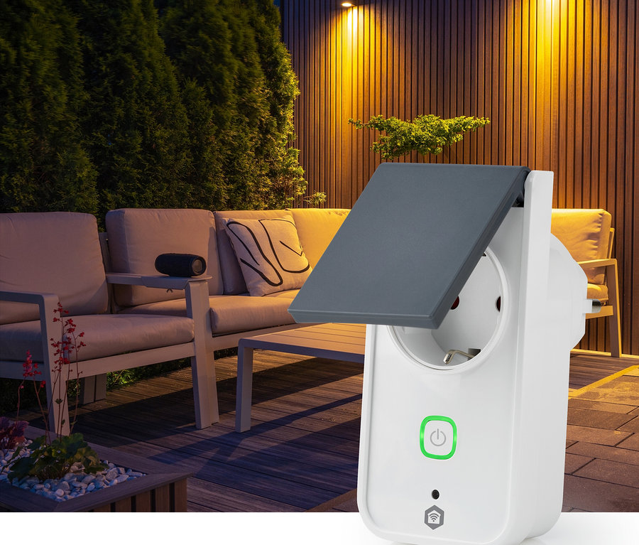 Ein weißer Smart Plug mit grauer, teilweise geöffneter Schutzabdeckung und grünem Einschaltknopf ist im Vordergrund. Dahinter eine unscharfe, nachts beleuchtete Außenterrasse mit warmer Wandbeleuchtung, Holzterrasse und hellen Gartenmöbeln.