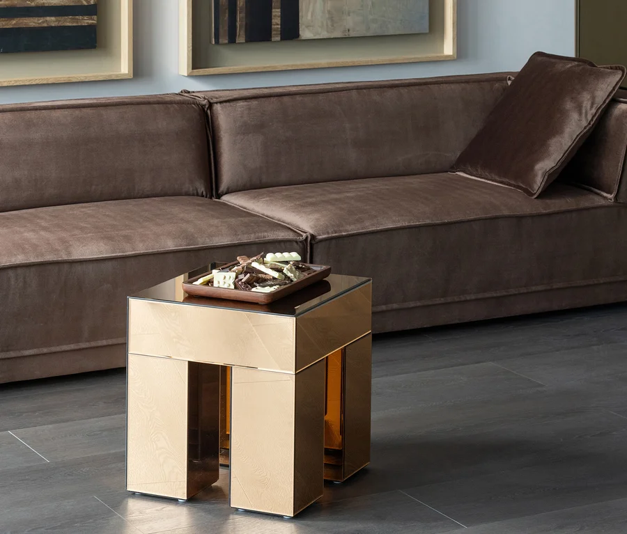 Goldener, spiegelnder Beistelltisch mit Holztablett voller Pralinen vor einem braunen Polstersofa mit passendem Kissen.
