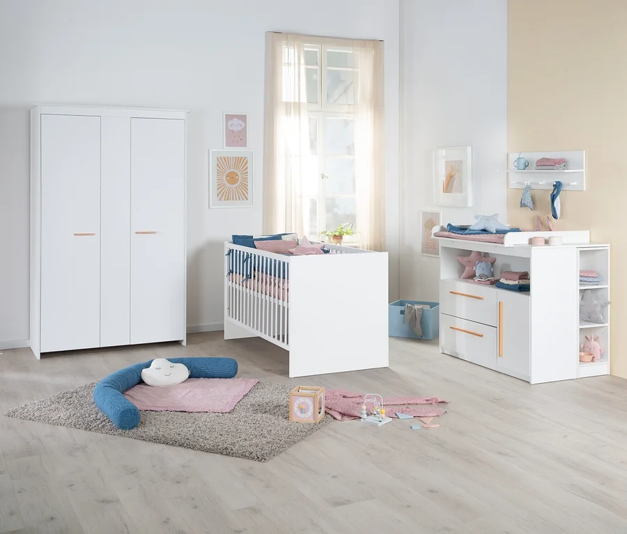 Helles Kinderzimmer mit weißen Möbeln: dreitüriger Kleiderschrank, Gitterbett mit bunter Bettwäsche und Wickelkommode mit Schubladen und offenen Fächern. Ein grauer Teppich liegt auf dem Boden mit Spielzeug.