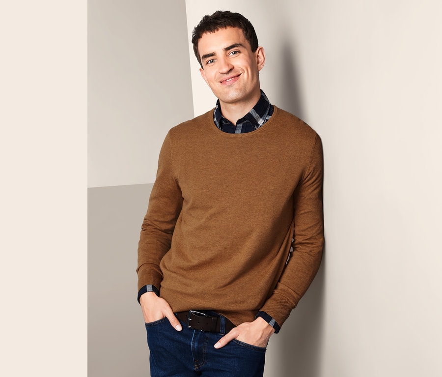 Mann lehnt an Wand, trägt braunen Pullover mit Rundhalsausschnitt und Jeans.