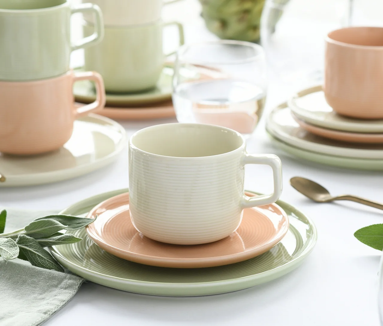 creaTable Service »Session Romance« auf einem Tisch, mit gerippten Tassen, Untertassen und Tellern in Pastellgrün, Rosa und Creme. Eine cremefarbene Tasse steht auf einer rosa Untertasse und einem grünen Teller im Vordergrund, weitere Teile sind gestapelt. Ein Wasserglas und ein goldener Löffel sind ebenfalls zu sehen.