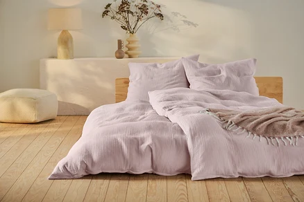 Rosafarbene Musselin-Bettwäsche in Normalgröße und Übergröße auf einem Bett mit Holzrahmen.