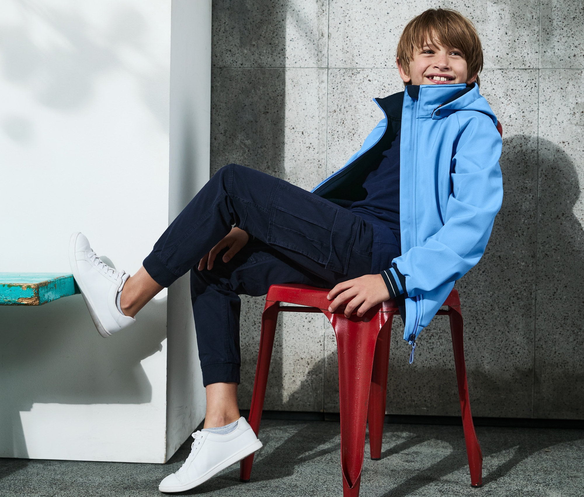 Ein Junge sitzt auf einem roten Stuhl und trägt eine Softshelljacke mit recyceltem Material.