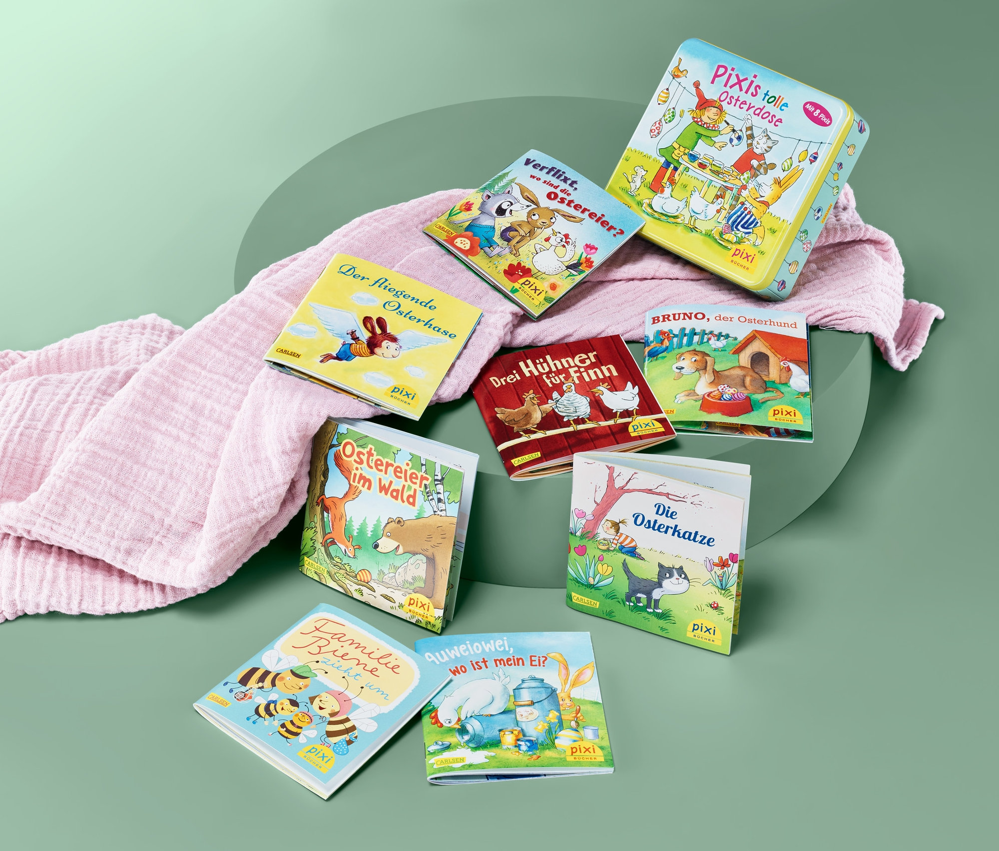 Eine Sammlung von Pixi-Kinderbüchern und Pixis tolle Osterdose liegen auf einem grünen Hintergrund und einem rosafarbenen Stoff.