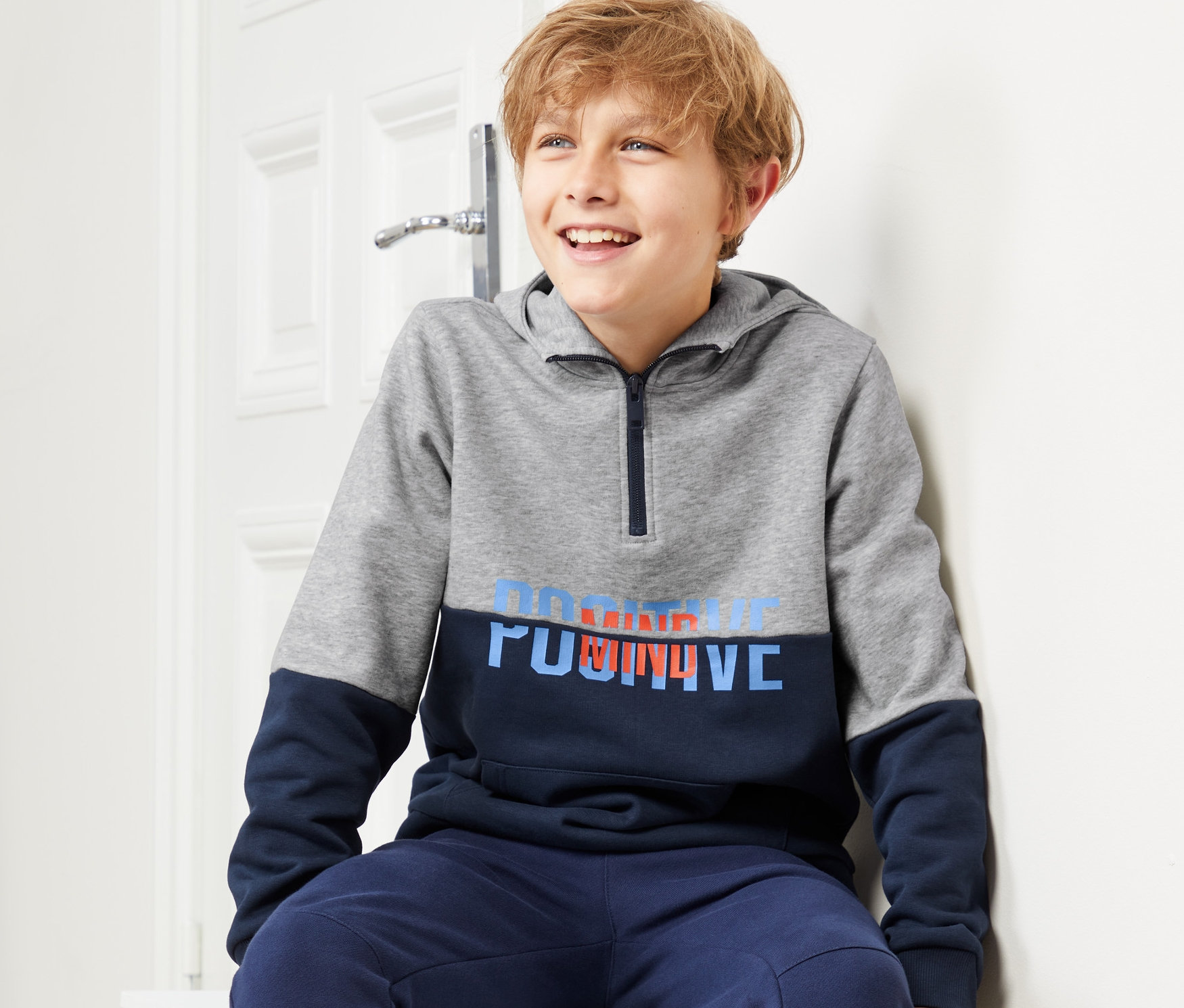 Ein Junge sitzt auf einer weißen Truhe und trägt einen grau-blauen Hoodie.