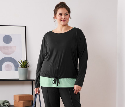 Frau steht barfuß da und hält eine Yogamatte. Sie trägt ein schwarz-grünes 2-in-1-Sportshirt und eine schwarze Sweathose.