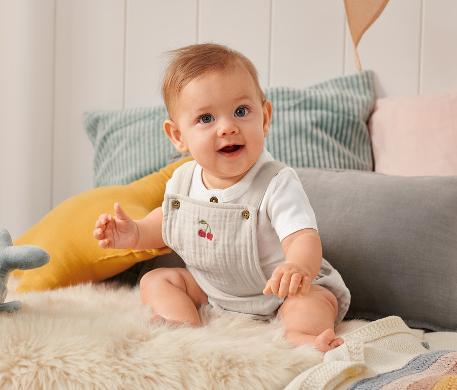 Lächelndes Baby sitzt auf einem Fell und trägt einen Baby-Strampler mit Kirschstickerei.