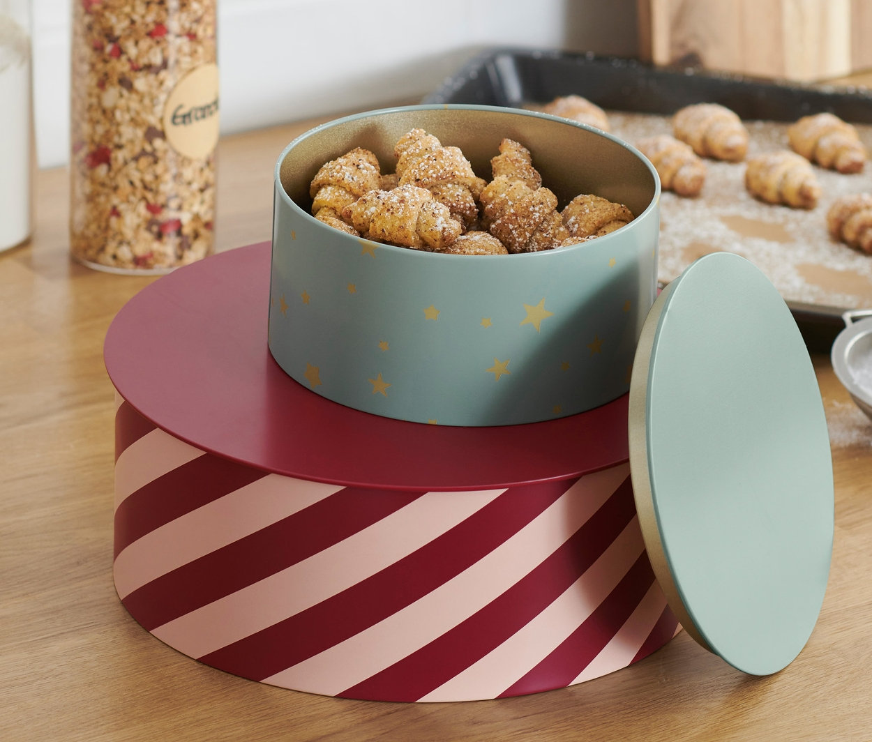 Zwei Keksdosen sind aufeinander gestapelt. Die obere Dose ist hellgrün mit goldenen Sternen und voller Mini-Croissants. Die untere Dose hat rot-rosa Streifen.