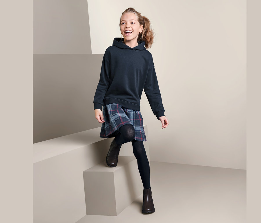 Lächelndes Mädchen sitzt auf Stufen und trägt ein marineblaues Sweatkleid und einen karierten Rock.