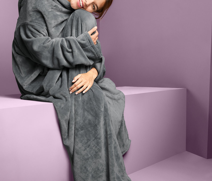 Frau sitzt auf einem lila Würfel, trägt eine Kuscheldecke mit integrierten Ärmeln und lächelt.