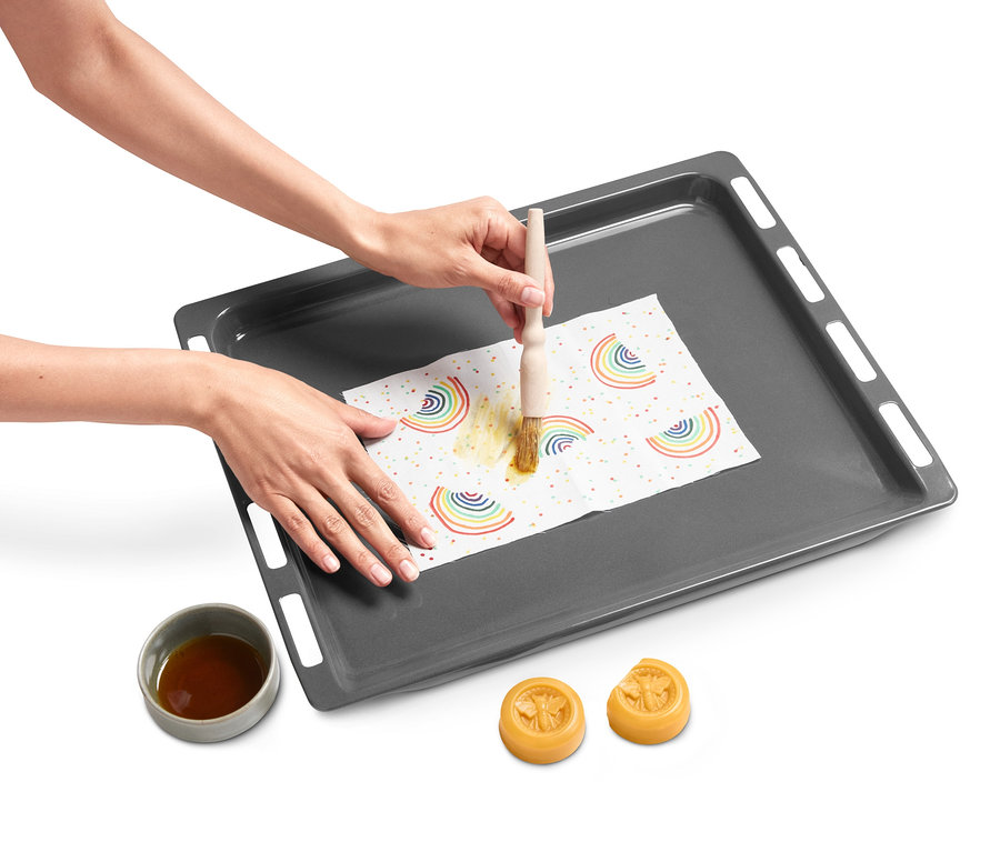 Frau pinselt Wachs auf ein Tuch mit Regenbogenmuster auf einem Backblech. Im Bild ist die Little Bee Fresh DIY-Box für Bienenwachstücher zu sehen.