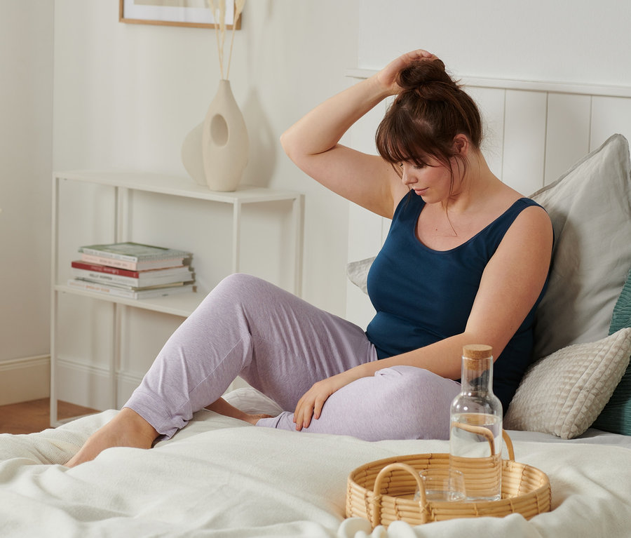 Frau sitzt im Bett mit einem der 2 Hemdchen und einer Relaxhose und macht sich einen Dutt. Auf dem Bett steht ein Tablett mit Wasser.