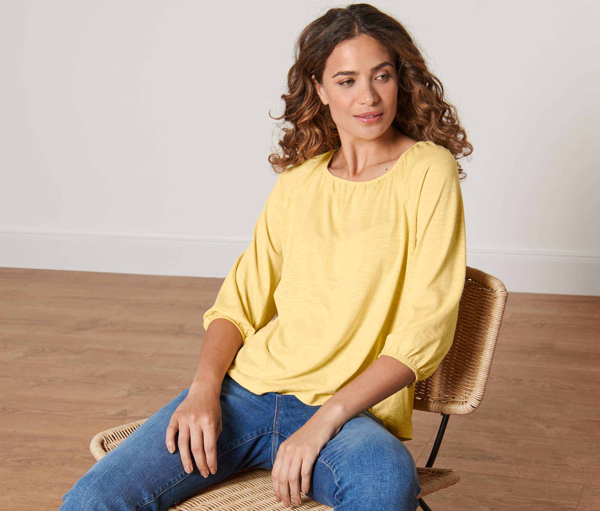 Eine Frau sitzt auf einem Rattanstuhl und trägt ein gelbes Blusenshirt mit 3/4-Arm.