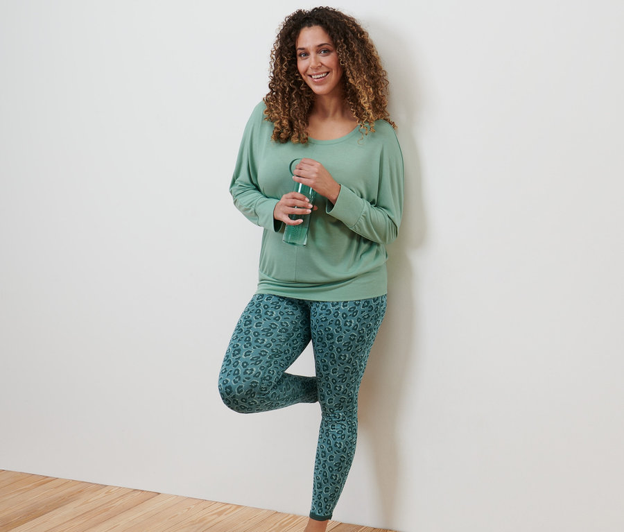 Eine Frau lehnt an einer Wand und trinkt Wasser. Sie trägt ein Sport-und-Yogashirt, salbeigrün und eine Wende-Sporttight, smaragd und salbei.