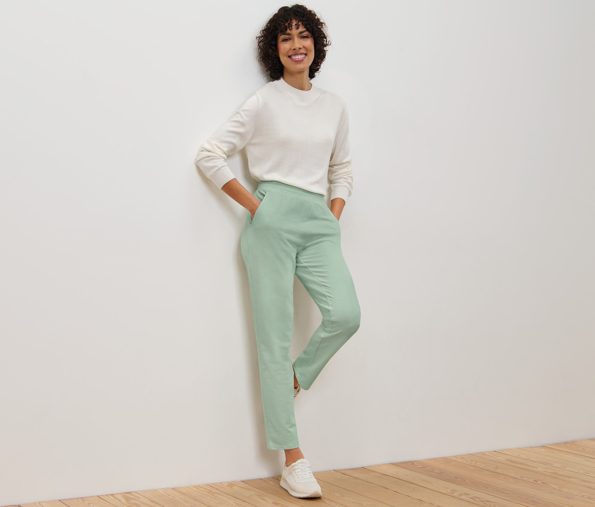 Frau lehnt an Wand, trägt Joggpants und Pullover.