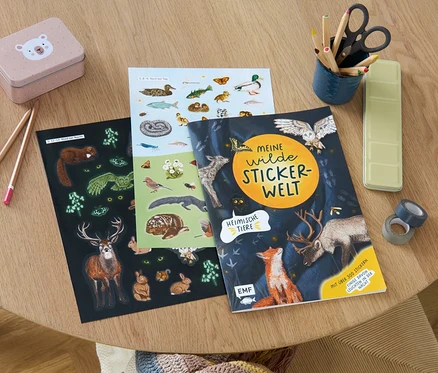 Auf einem Holztisch liegen Stifte, eine Schere, eine Dose und das Stickerbuch »Meine wilde Stickerwelt – heimische Tiere«.
