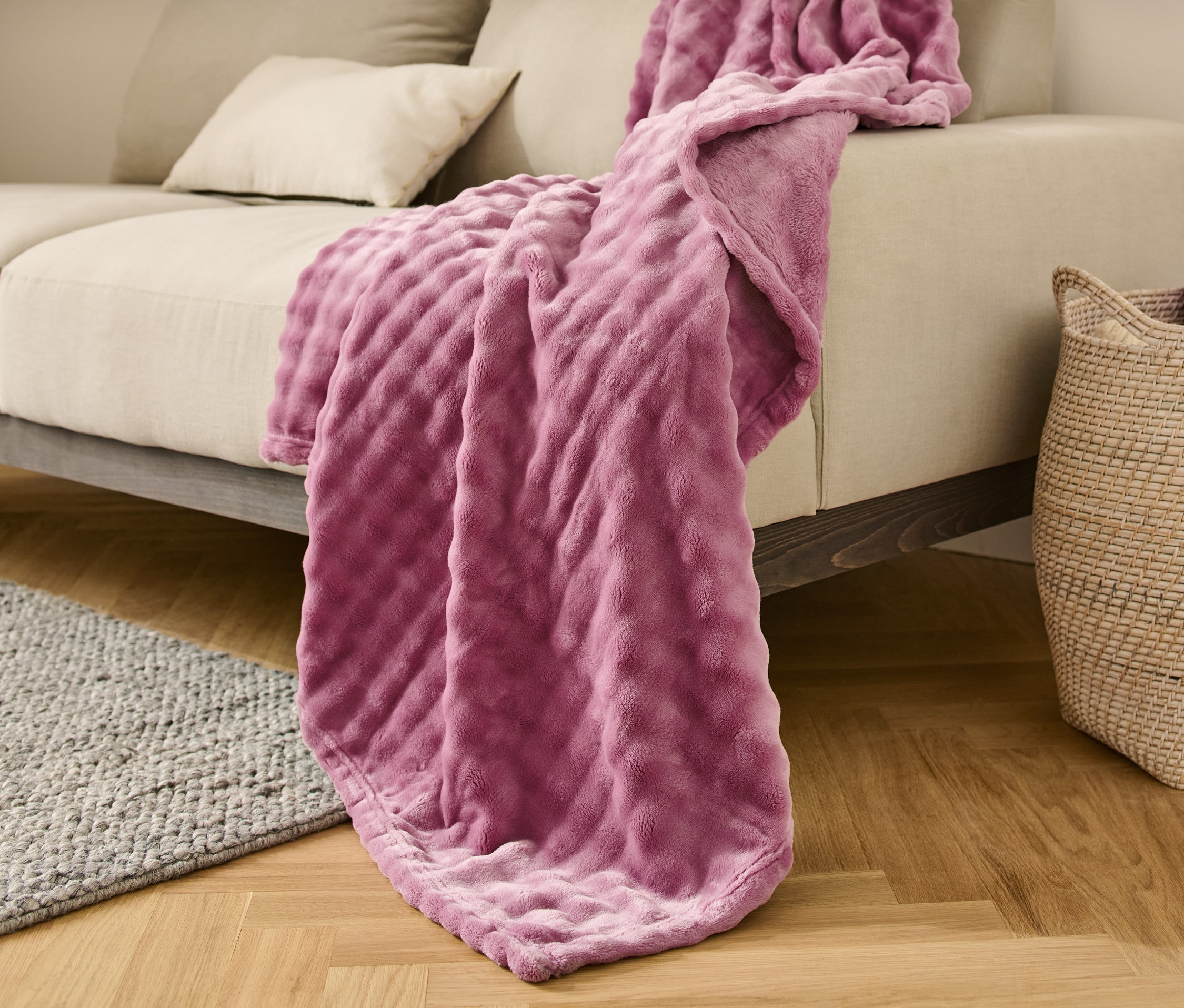 Eine Kuscheldecke, ca. 150 x 200 cm, liegt auf einem beigen Sofa. Neben dem Sofa steht ein Weidenkorb.