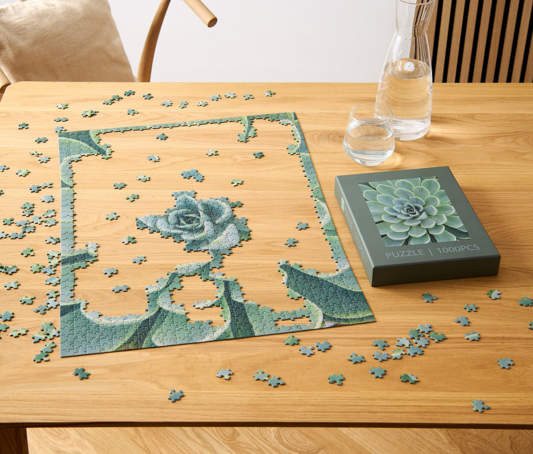 Ein teilweise zusammengesetztes Puzzle »Sukkulente« liegt auf einem Holztisch neben einer Schachtel mit Sukkulentenmotiv, einem Glas und einer Karaffe Wasser.