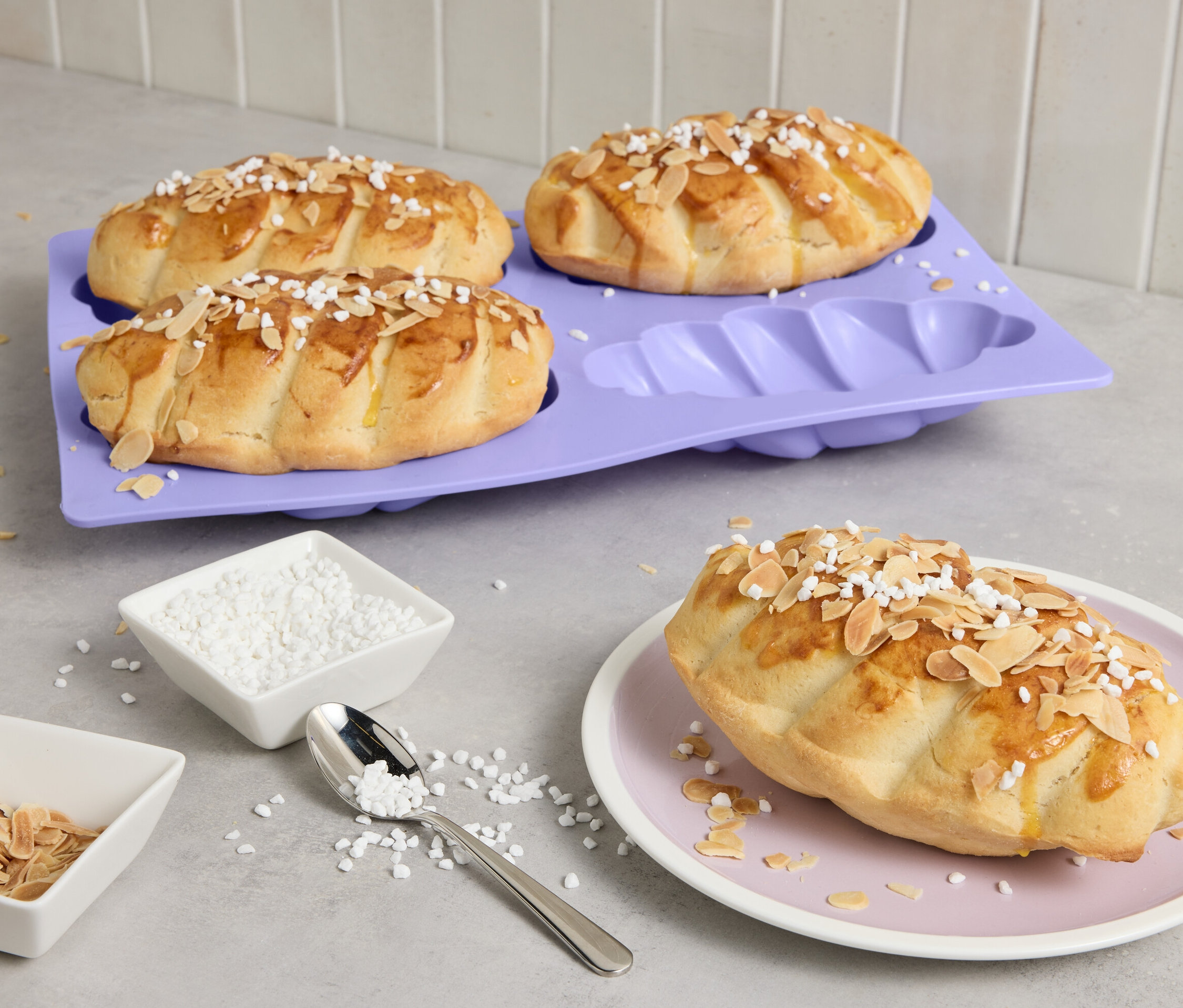 Vier Mini-Hefezöpfe auf lila Backform, einer auf Teller, mit Mandeln und Zucker bestreut.