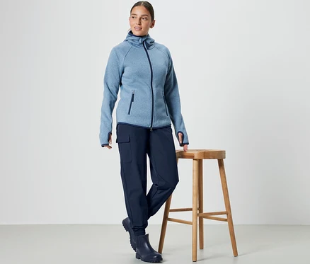 Eine Frau steht neben einem Holzhocker und trägt eine blaue Kapuzenjacke und eine Softshell-Joggpants.