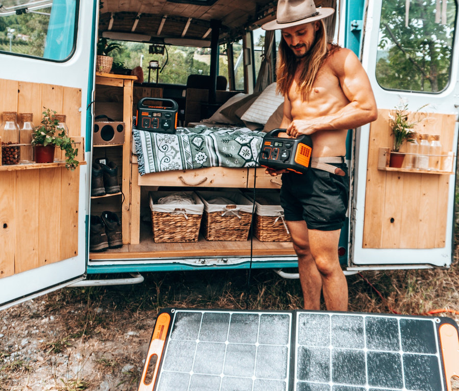 Mann mit Hut hantiert mit Jackery Explorer 240 vor Van, Solarmodul SolarSaga 100 liegt am Boden.