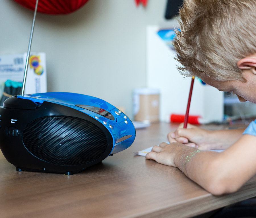 Junge schreibt auf Papier neben blauem Lenco »SCD-24 Boombox« CD-Player mit Radio.