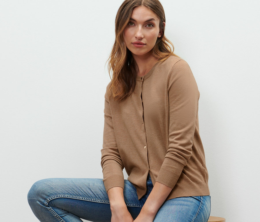 Eine Frau sitzt auf einem Holzhocker und trägt einen Feinstrick-Cardigan, Jeans und Turnschuhe.